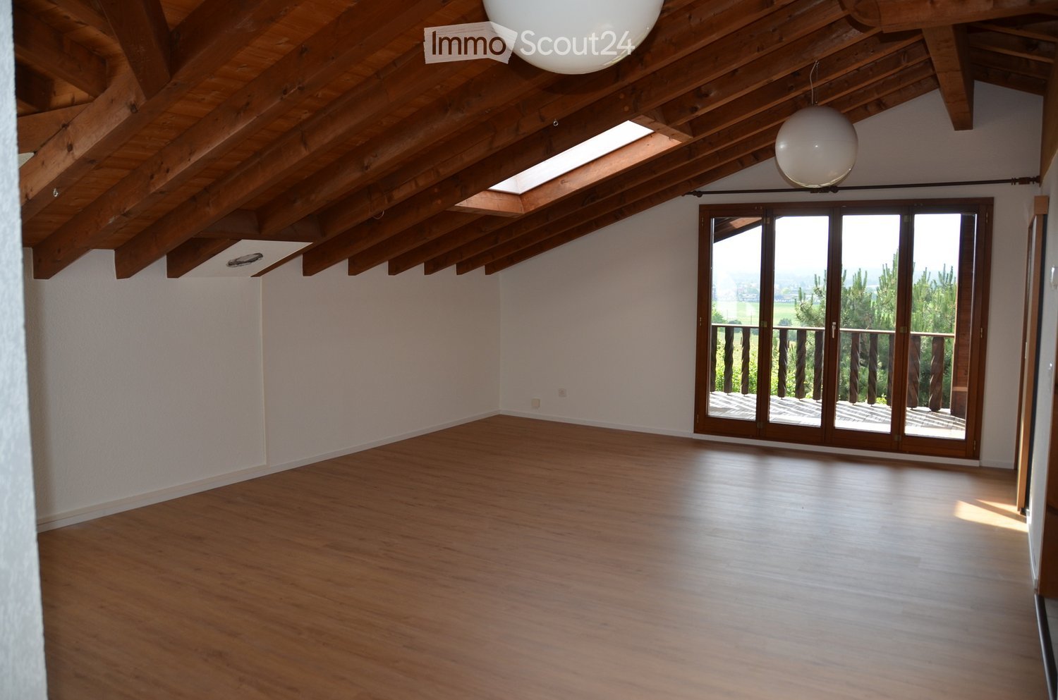 An empty room with wooden flooring, white walls, and a wooden ceiling with exposed beams. A large sliding glass door leads to a balcony with a scenic view.