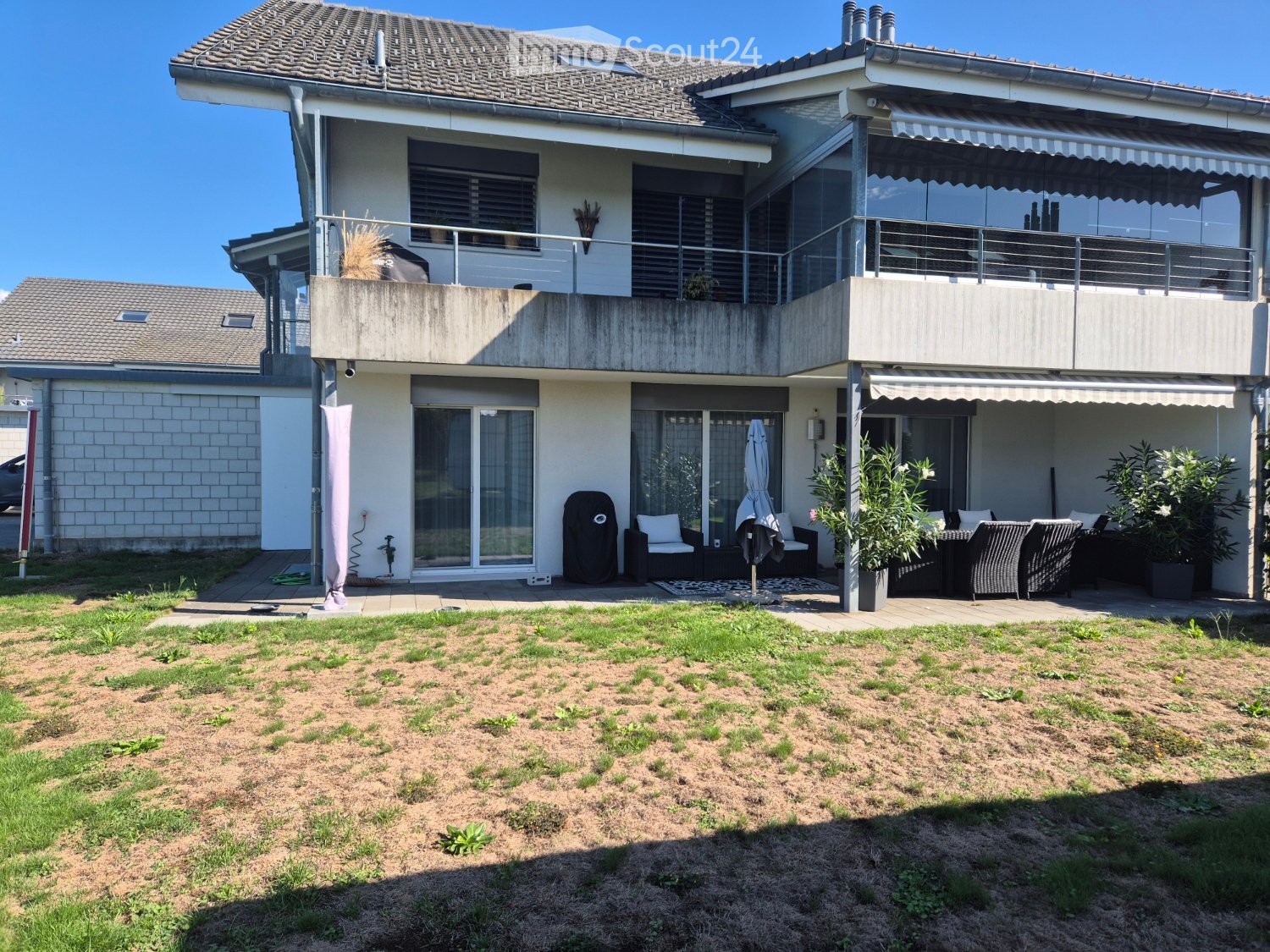 Two-story house with a modern design, featuring a large balcony, patio seating area, and a well-maintained lawn.