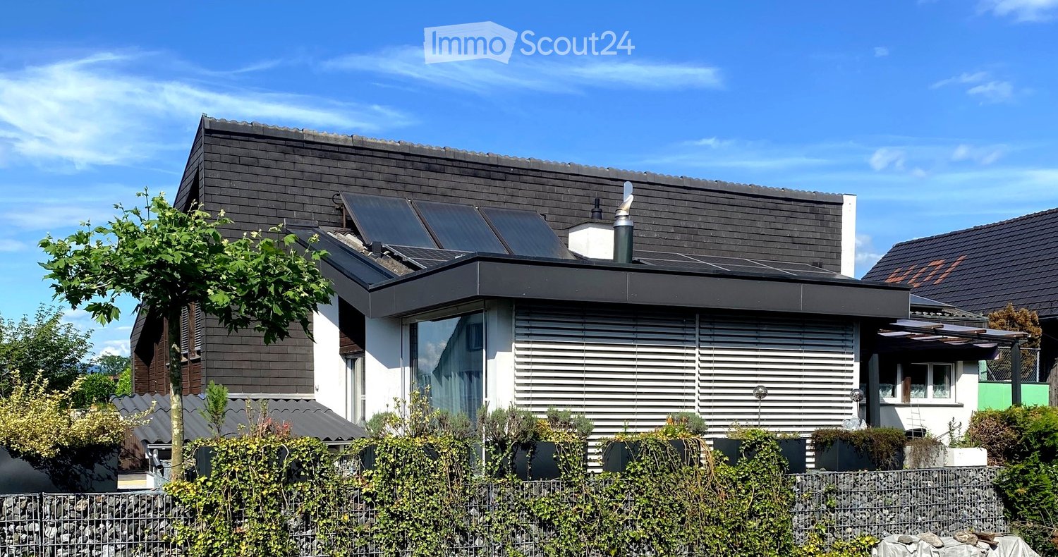 Modern residential building with a flat roof, solar panels, and a small outdoor seating area. The structure features large windows and a sleek, contemporary design.