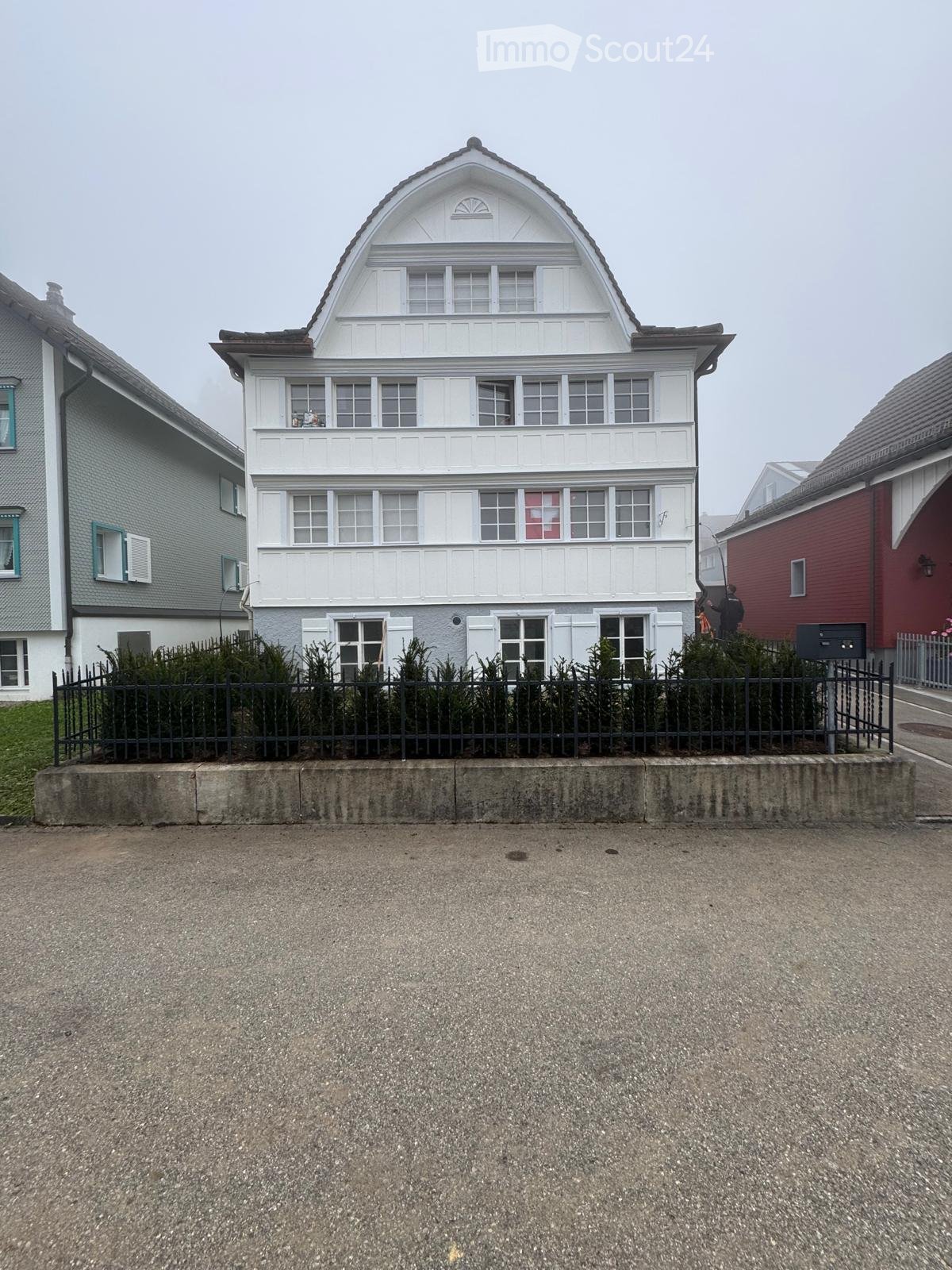 Building with several apartments, white exterior, multiple windows, gray neighboring houses, black fencing, grassy area, road