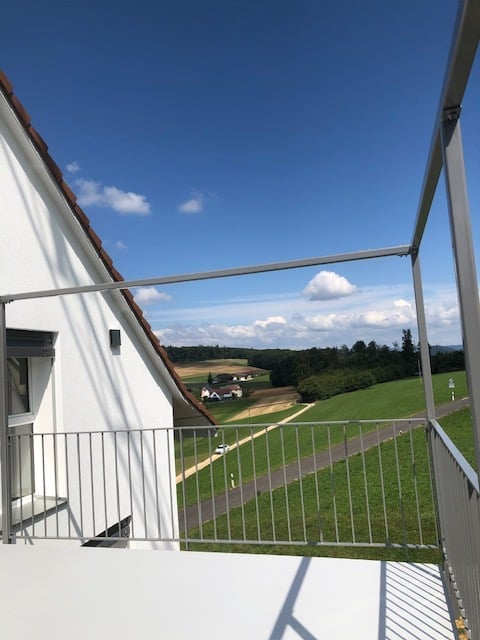 Balcony with metal railing, overlooking a green landscape with fields and a road.