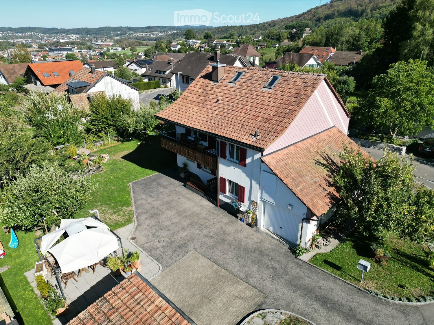 Aerial view of a two-story house with a red-tiled roof, white exterior, and a balcony. The property includes a driveway, a small garden area with outdoor furniture, and is surrounded by other houses and greenery.