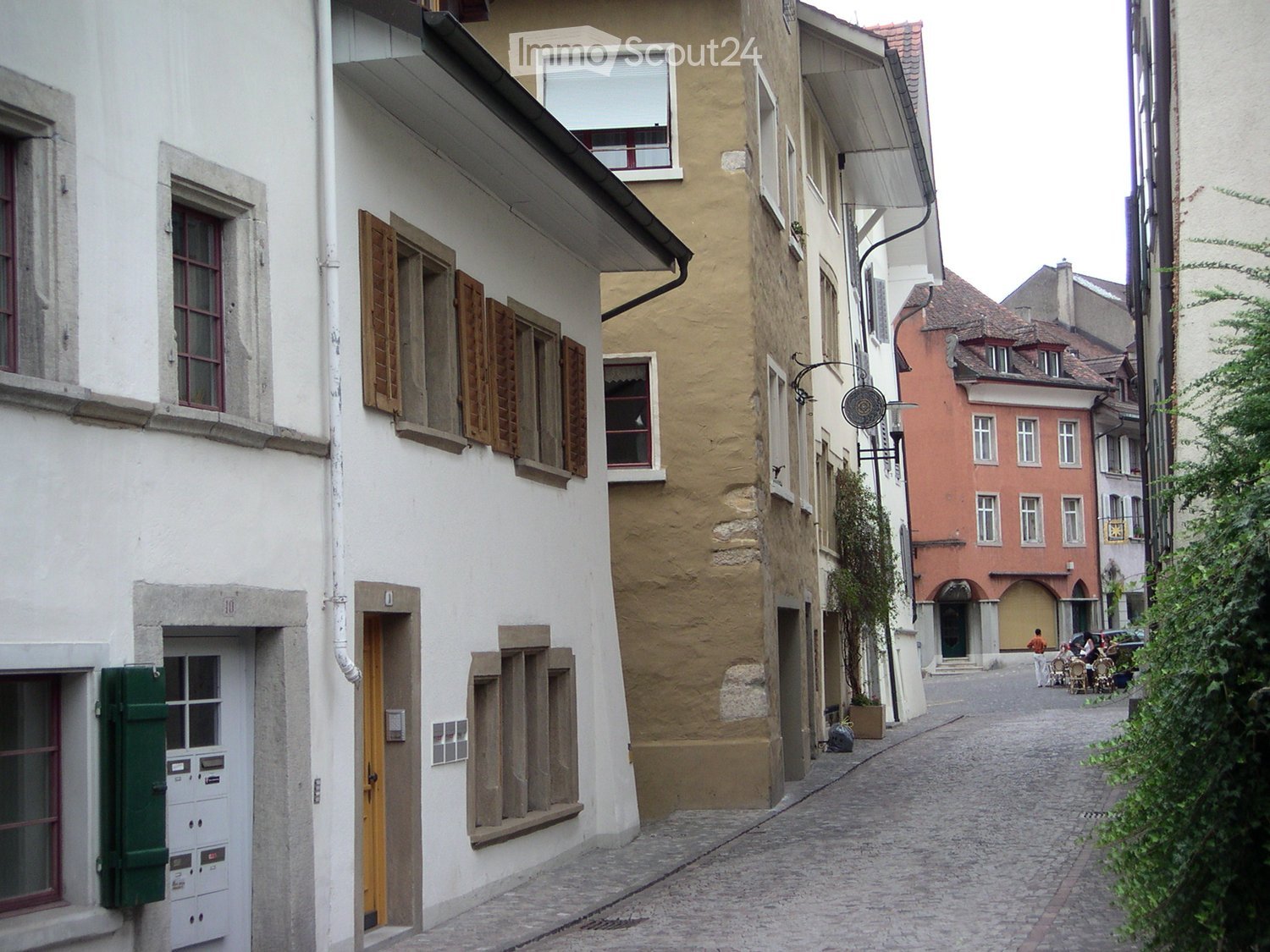 A narrow cobblestone street lined with multi-story buildings featuring shutters, windows, and doors. The architecture includes a mix of white and beige facades with traditional European design elements.