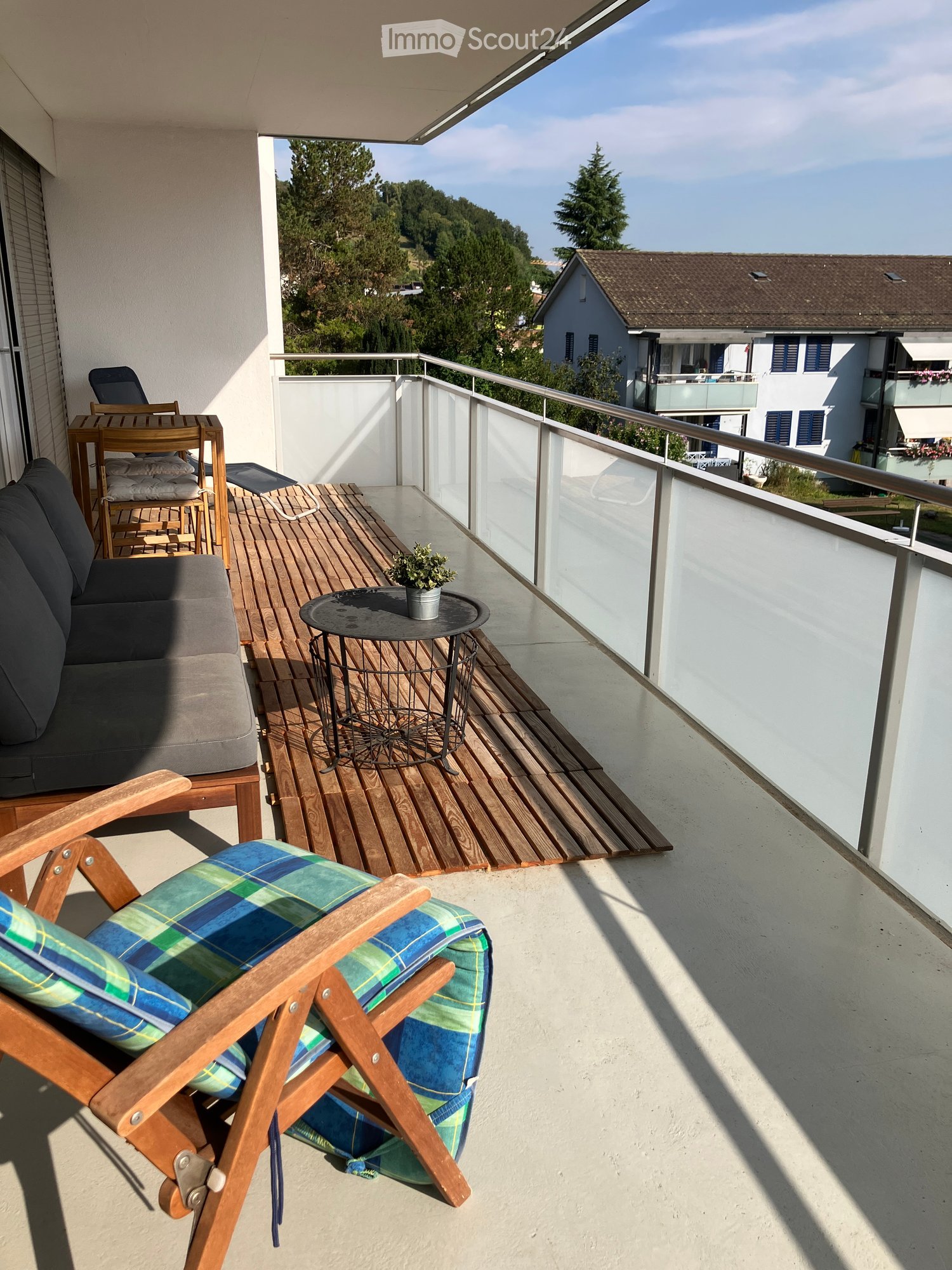 Balcony with wooden decking, sofa, small table with plant, wooden chairs, and a view of the surrounding area.