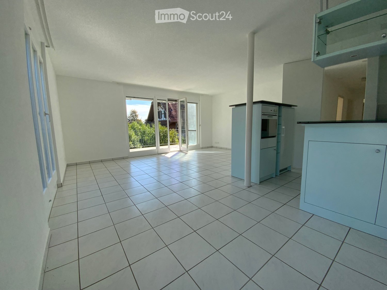 Empty room with tiled floor, white walls, large windows, and a small kitchen area with cabinets and appliances.