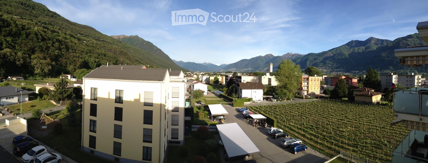 A multi-story residential building with a parking lot, surrounded by vineyards and a scenic mountain view.