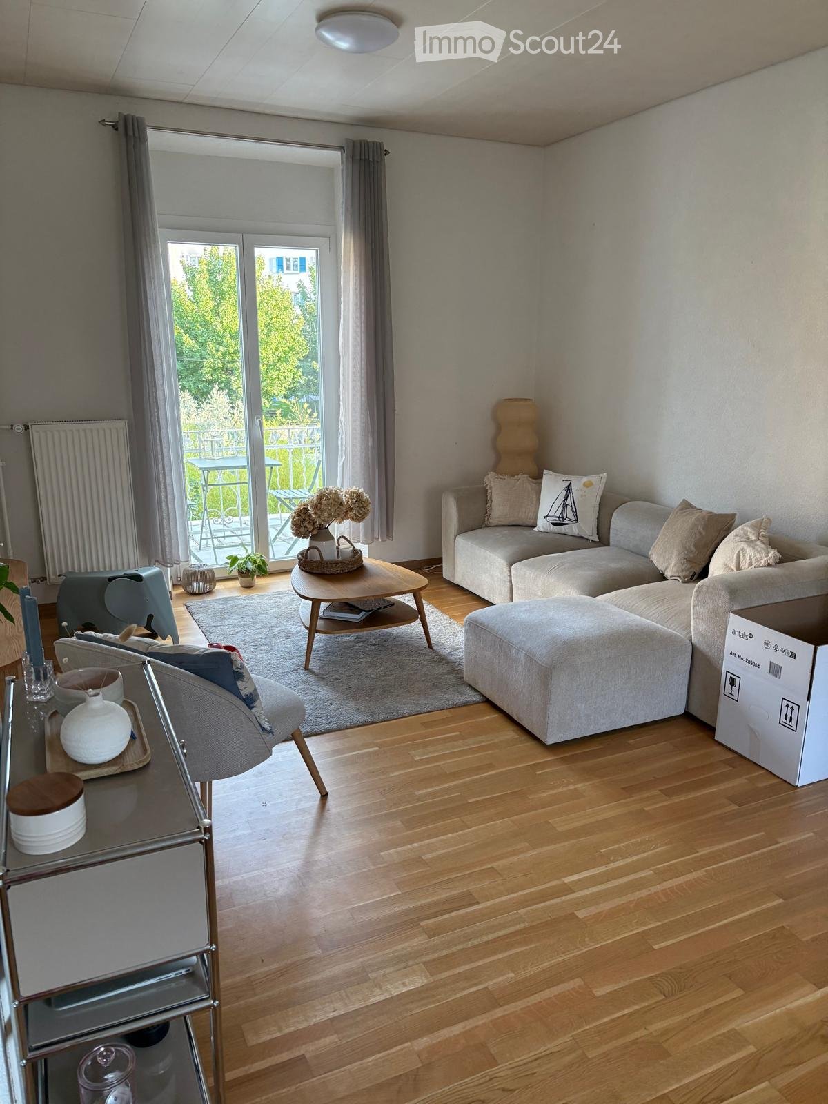 Modern living room with light wooden flooring, grey sectional sofa, coffee table, ottoman, and a small dining table leading to a balcony.