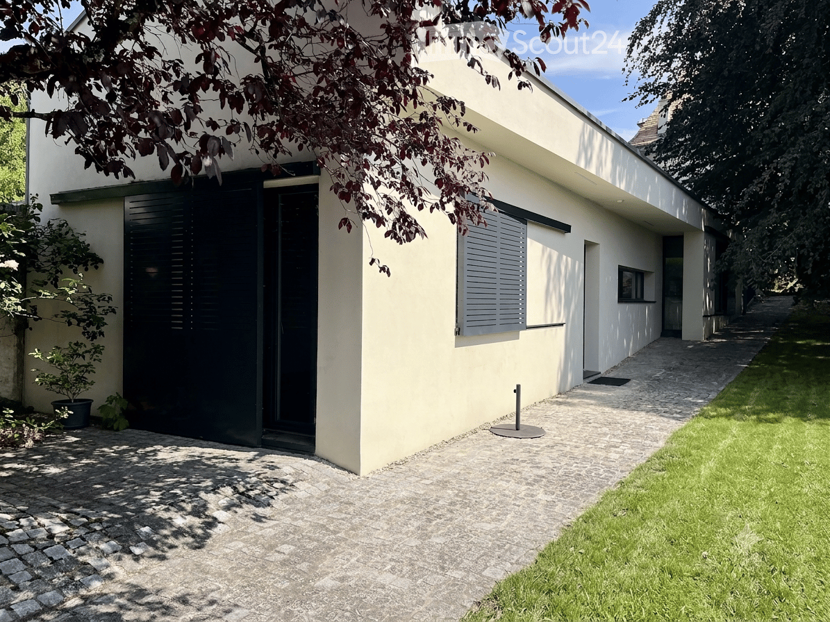 Modern single-story house with light yellow walls, black shutters, and a paved entrance path. A small potted plant is near the entrance, and the house is partially shaded by tree branches.
