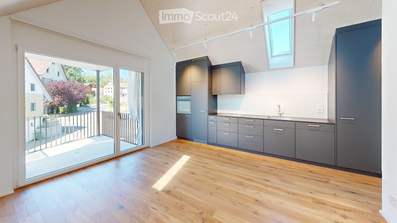 Modern kitchen with grey cabinets, wooden flooring, and large windows overlooking a balcony.