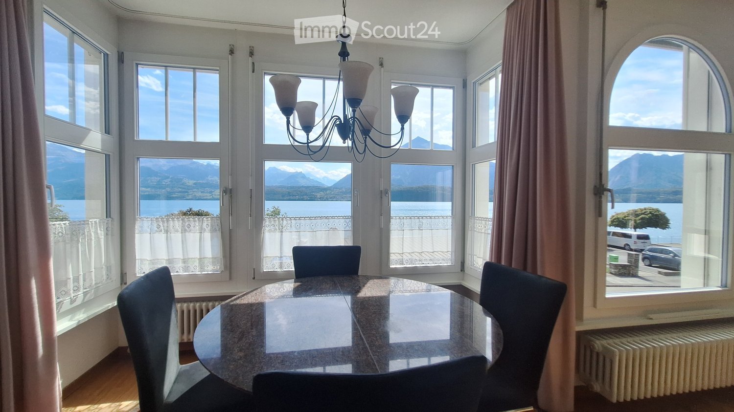 Bright dining room with large windows offering a scenic mountain and lake view, featuring a round glass dining table, four chairs, and a chandelier.