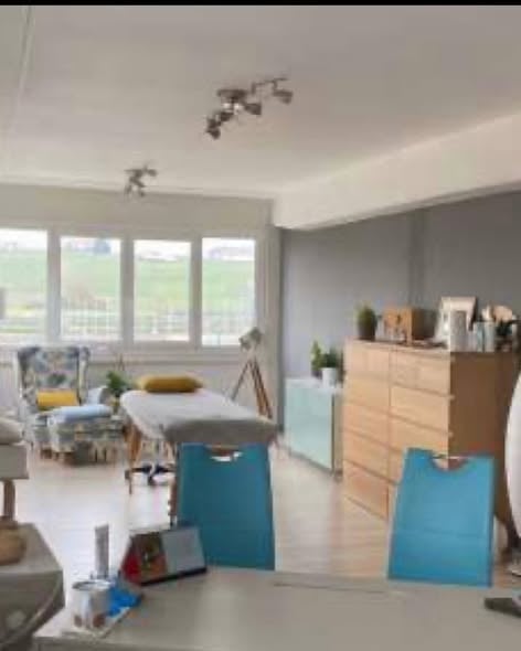 Bright, modern room with large windows, a sofa, a chair, a small table, a lamp, a mirror, and a cabinet with decorative items.