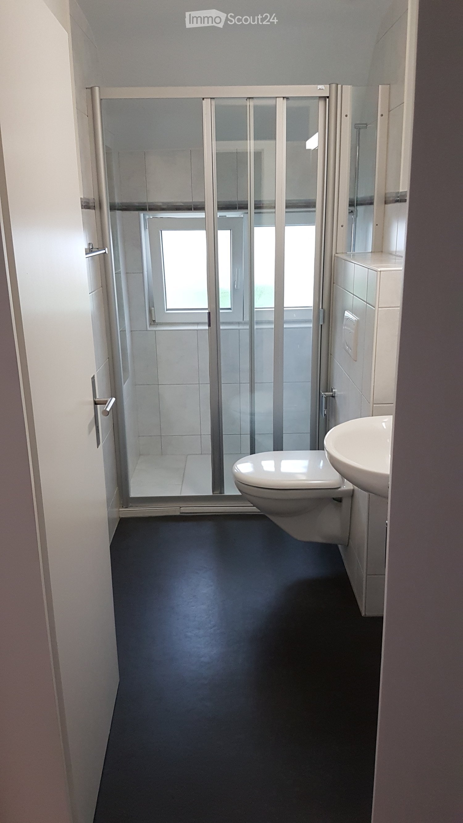 Modern bathroom with dark flooring, white walls, a toilet, a sink, and a glass shower enclosure with a window.