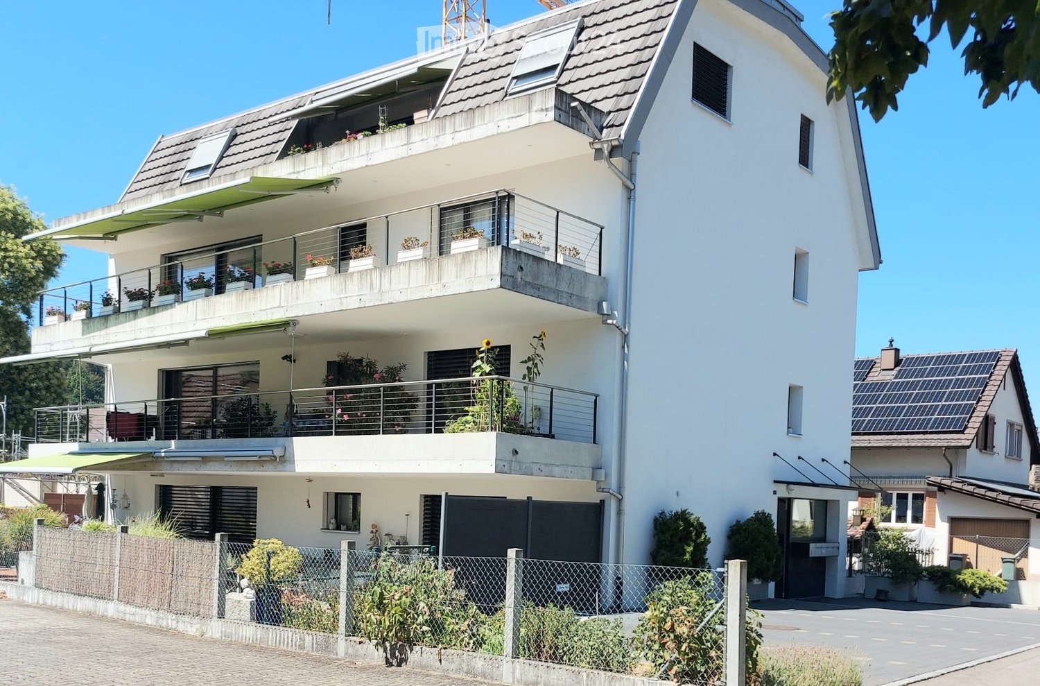 Modern two-story house with white exterior, multiple balconies, and a garage. Features include large windows, a covered balcony area, and solar panels on an adjacent building.