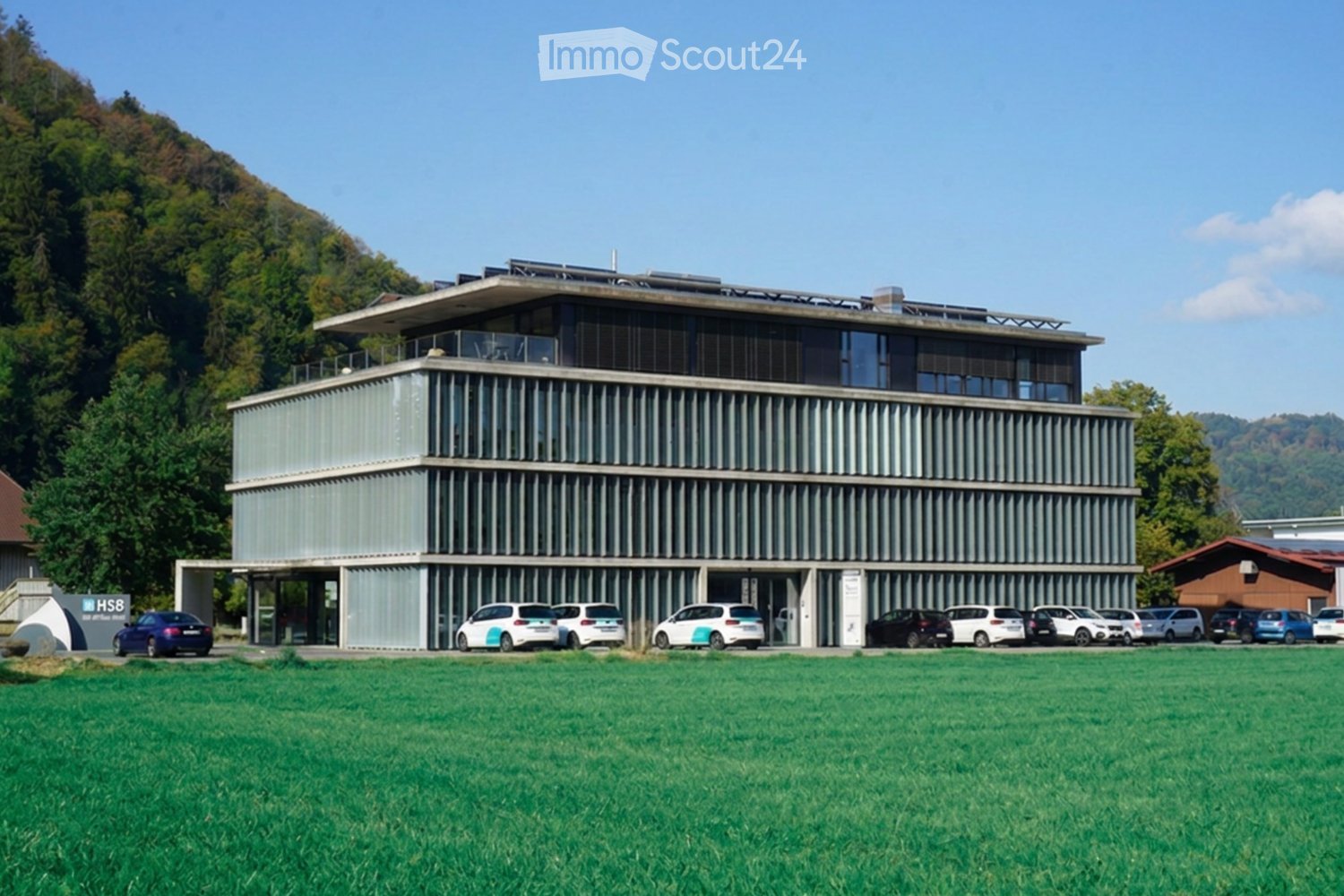 Modern two-story building with a sleek, contemporary design, featuring large windows and a flat roof, surrounded by a grassy field and parked cars.