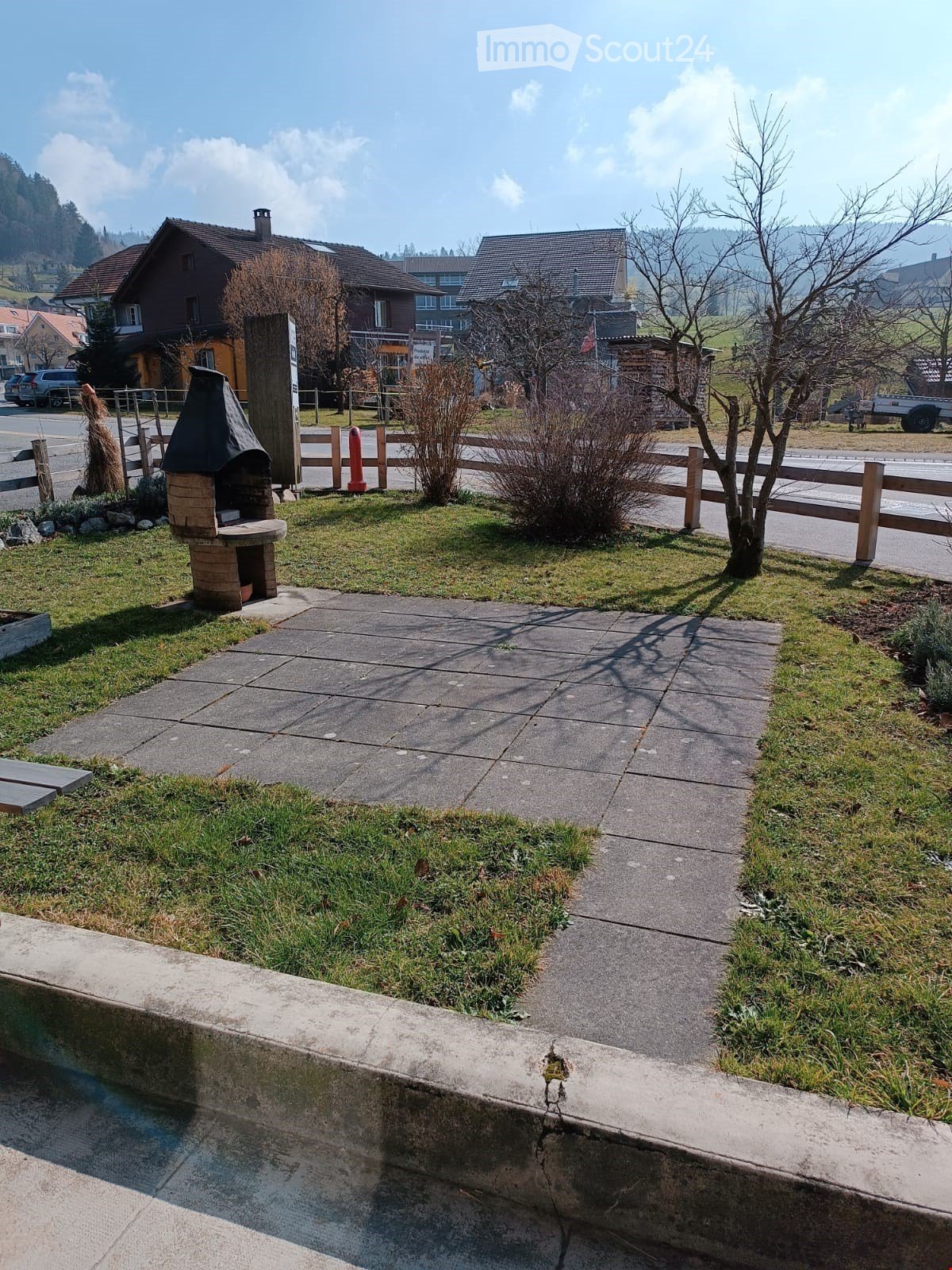 Outdoor area with a brick barbecue, wooden fence, small tree, and paved pathway.