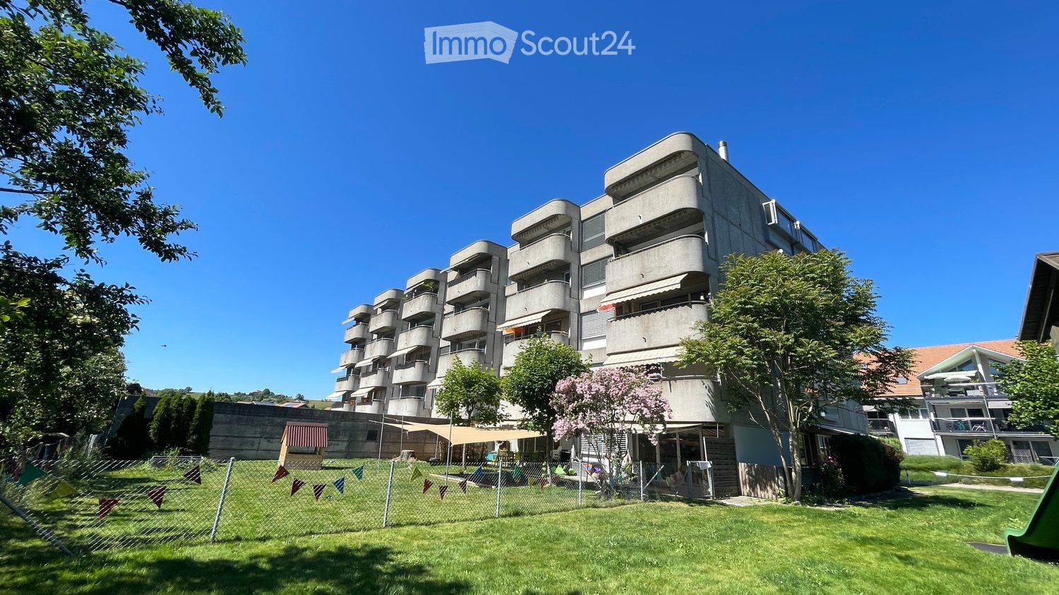 Modern multi-story apartment building with balconies, surrounded by greenery and a fenced yard with playground equipment.