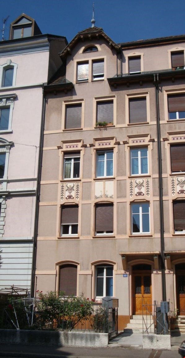 A multi-story residential building with beige and light pink facade, multiple windows with shutters, decorative moldings, and a small garden area in front.