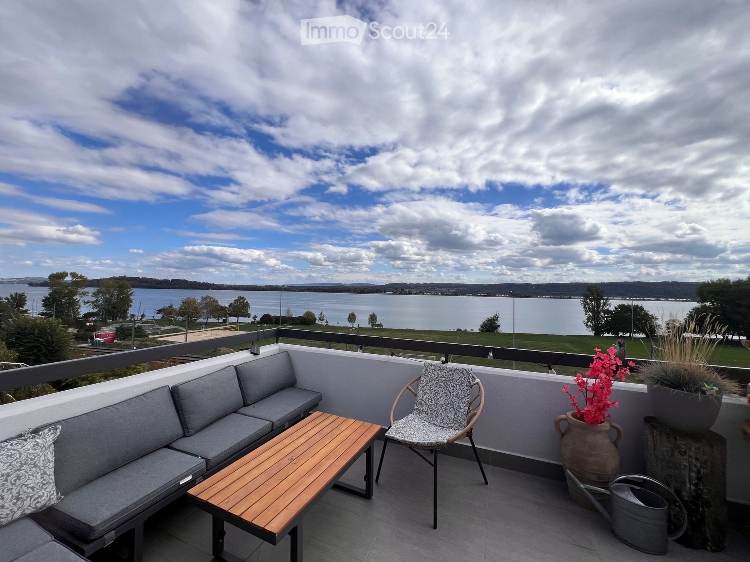 Balcony with grey sofa, wooden table, patterned chair, and flower pot overlooking a lake.