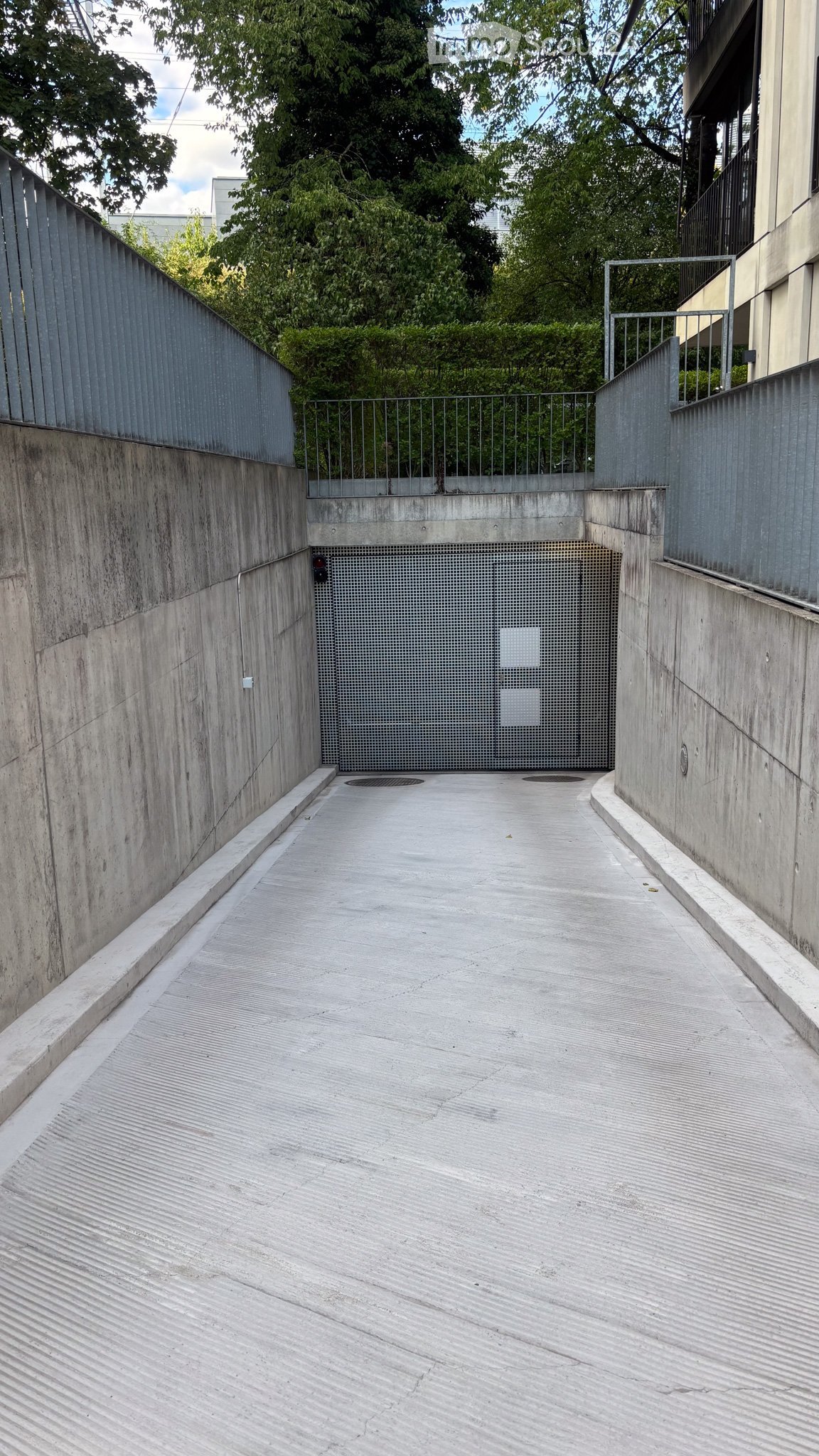 A narrow concrete corridor with metal railings on both sides, leading to a gated area with greenery visible beyond.