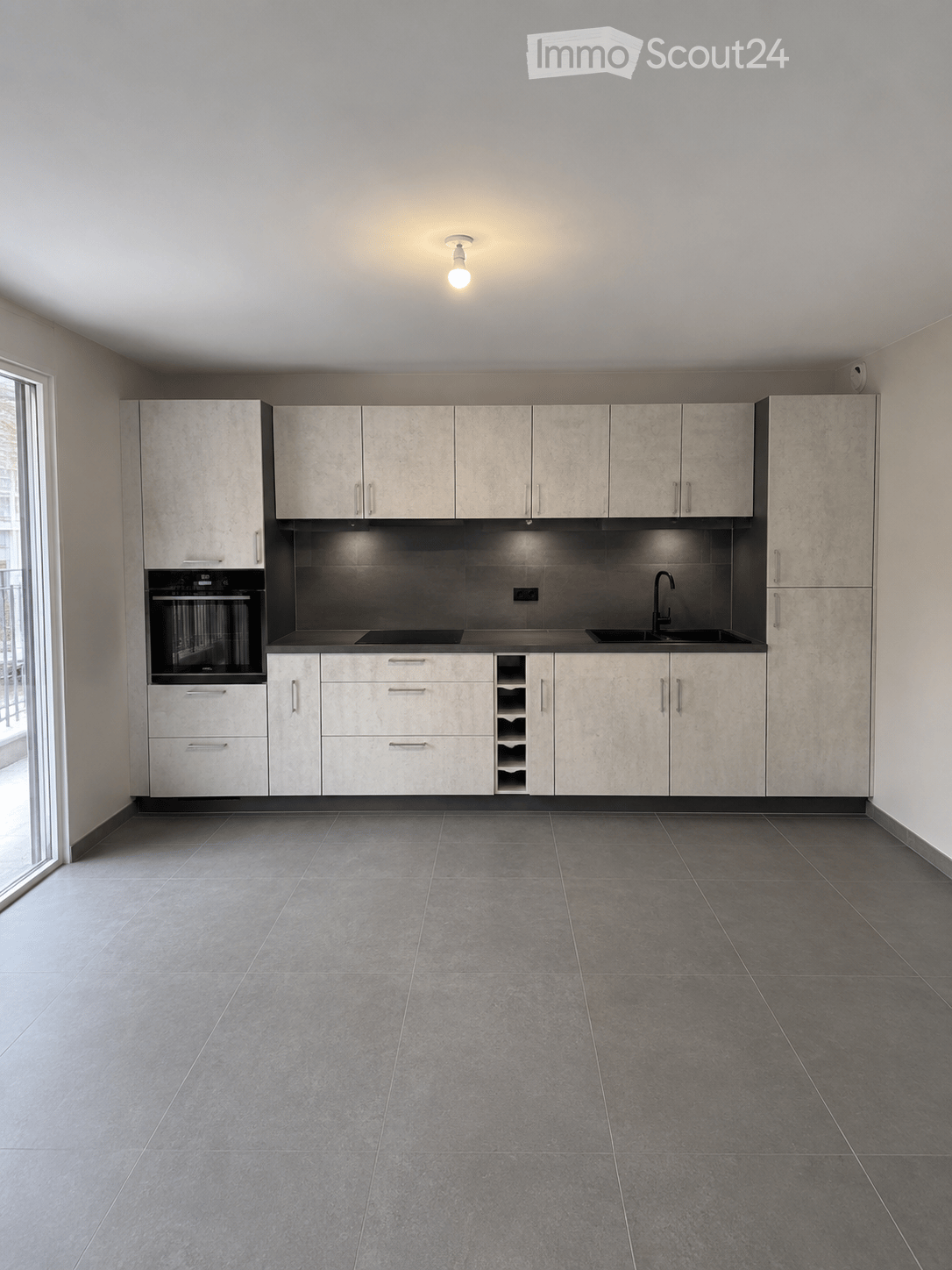 Empty kitchen with modern cabinets, stainless steel sink, and built-in oven. Grey tiled floor and white walls.