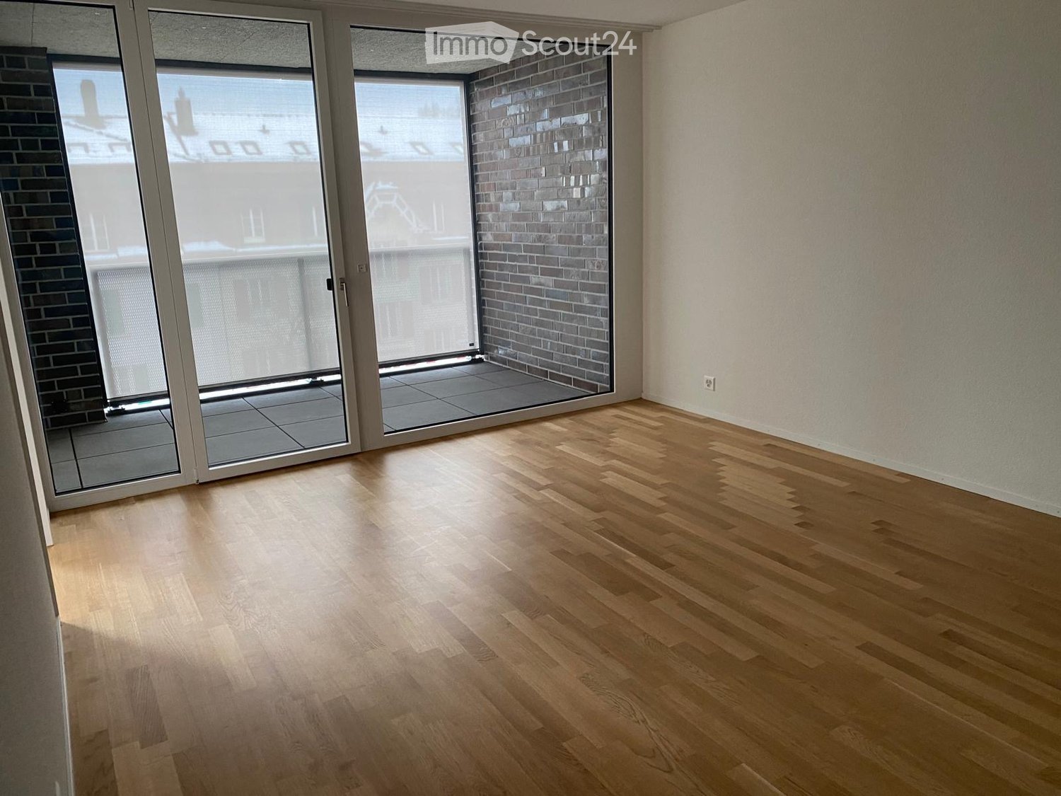Empty room with light wooden flooring, large sliding glass doors leading to a balcony, and white walls.