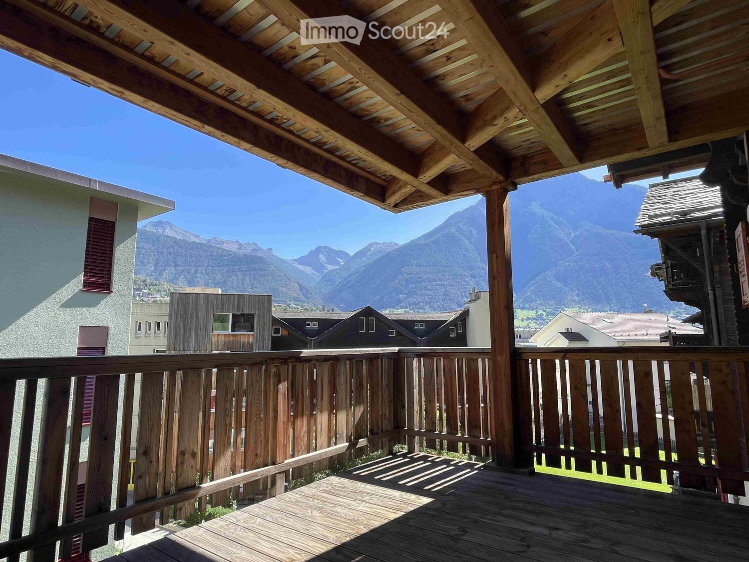 Wooden balcony with a railing, overlooking a scenic mountain view and neighboring houses.