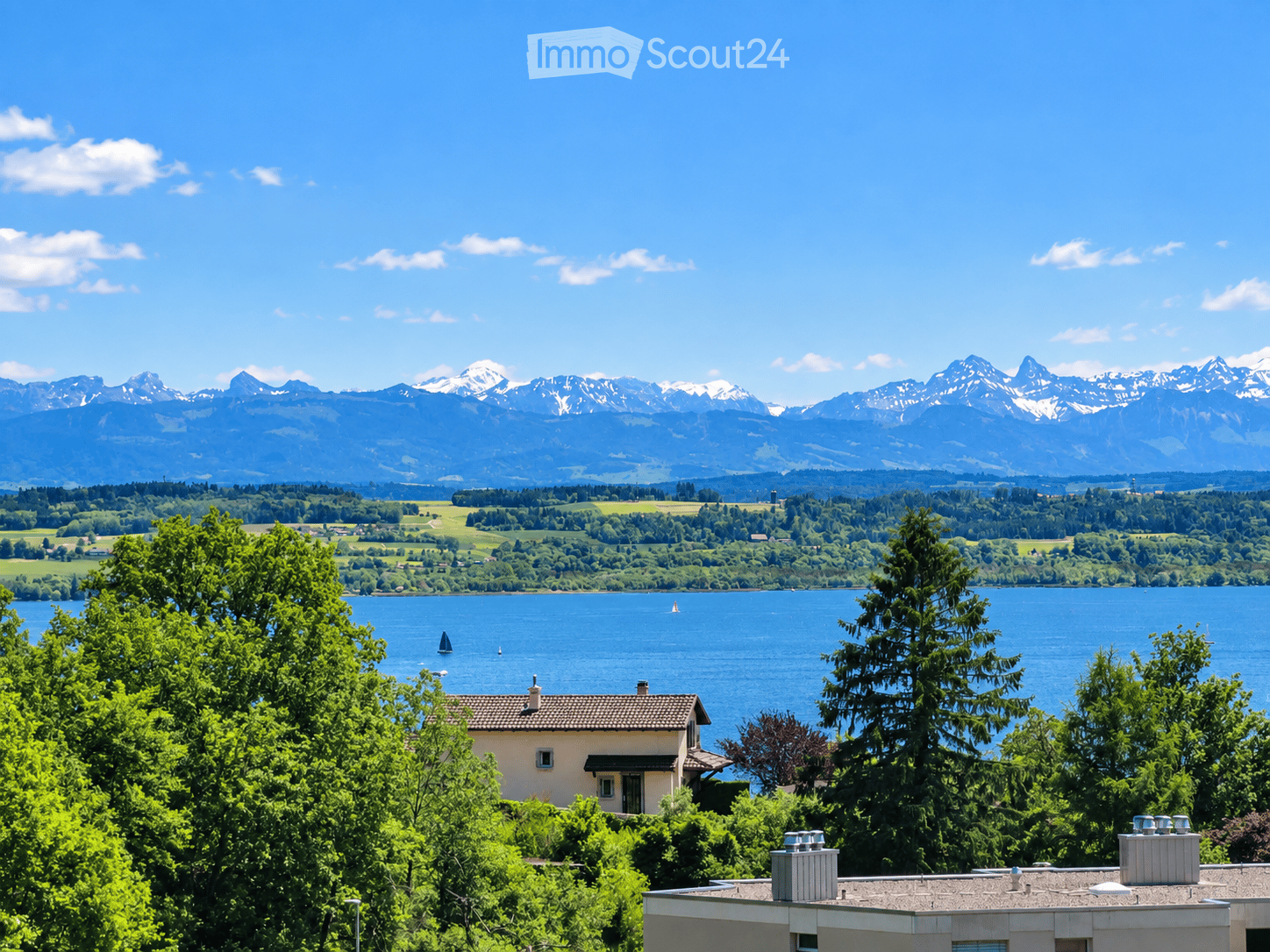 A picturesque view of a house nestled among lush greenery, overlooking a serene lake with sailboats, surrounded by rolling hills and snow-capped mountains under a clear blue sky.