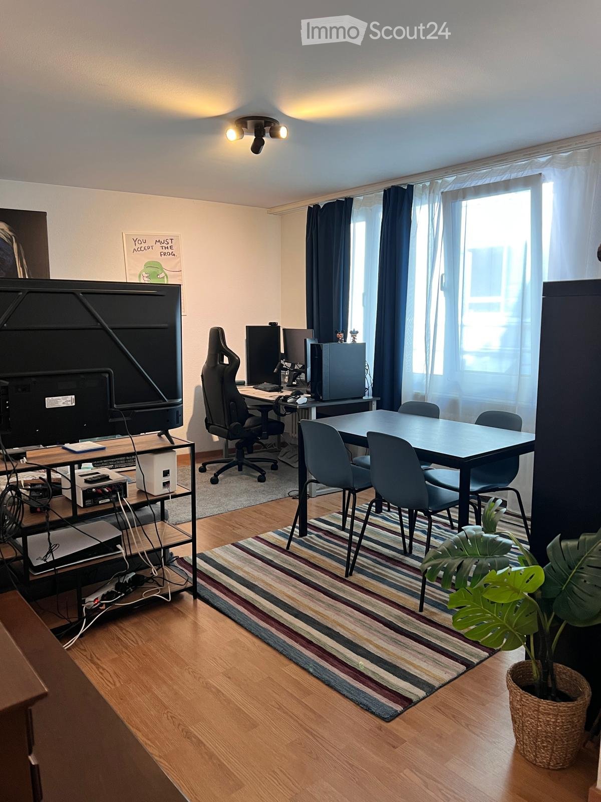 A modern, well-lit room with wooden flooring, a large window with curtains, a striped rug, a gaming chair, a desk with multiple screens, and a small table with chairs.