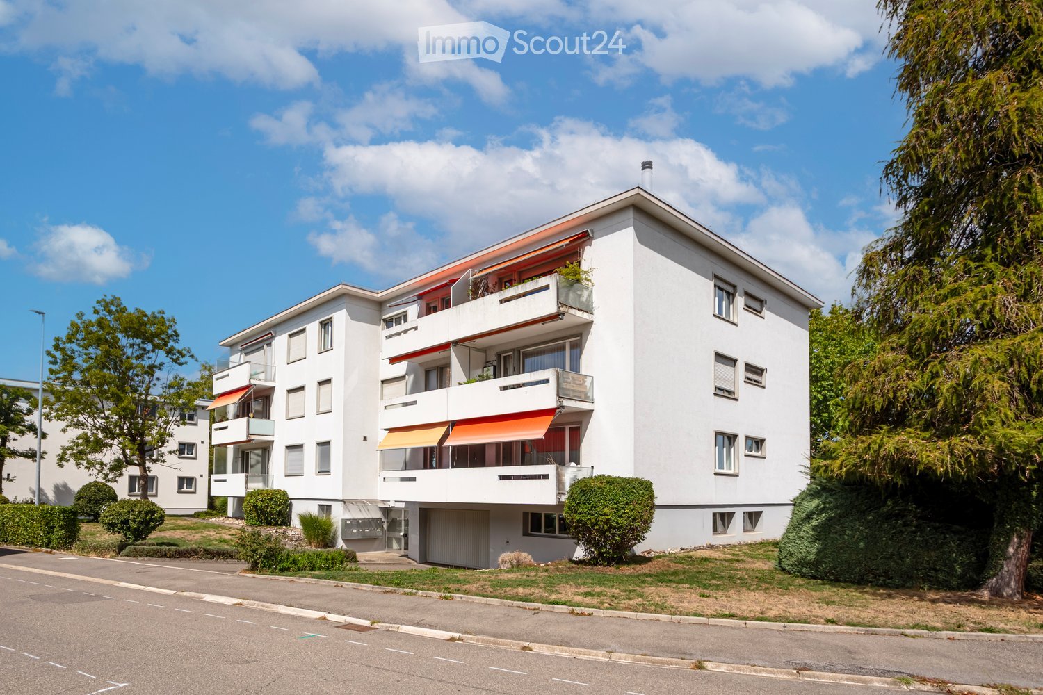 A modern, white multi-story apartment building with balconies, large windows, and orange awnings. The building is surrounded by greenery and trees, with a paved road in front.