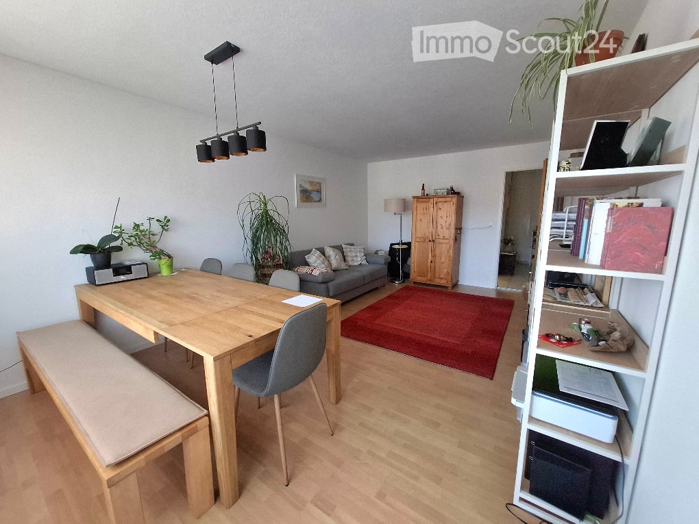 Bright, open-plan living and dining area with light wood flooring, a grey sofa, a wooden dining table with chairs, a wooden bench, a wooden cabinet, a bookshelf, and several potted plants.