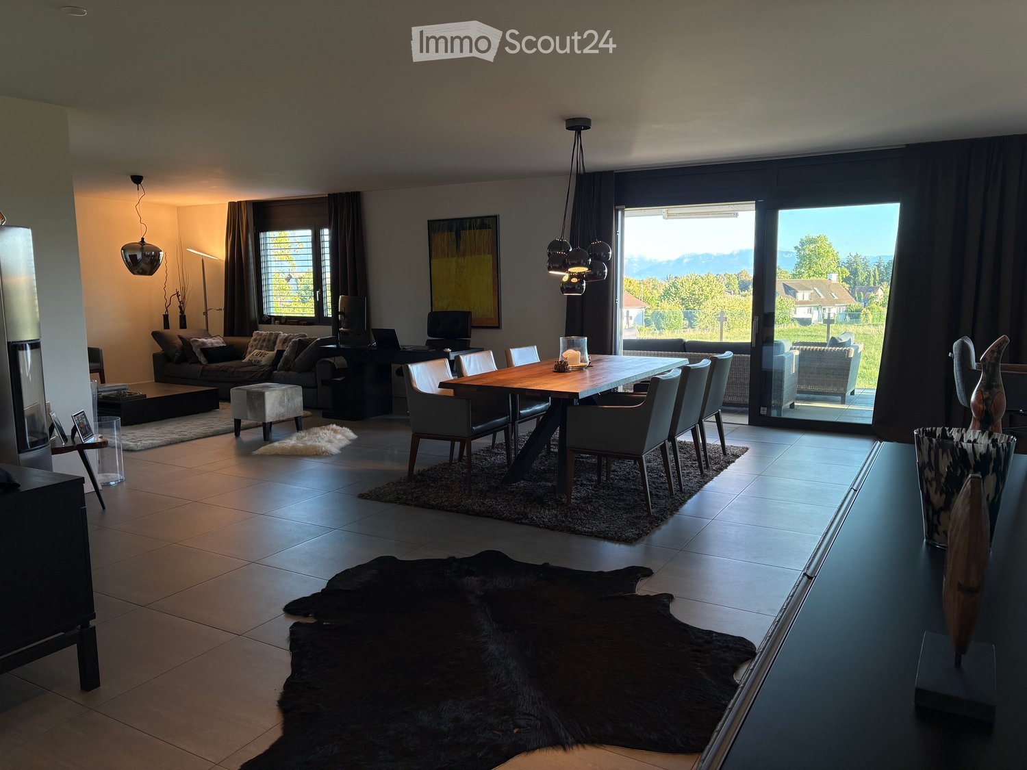 Modern living and dining area with large windows offering a scenic view, featuring a sofa, coffee table, ottoman, dining table with chairs, and decorative vases.