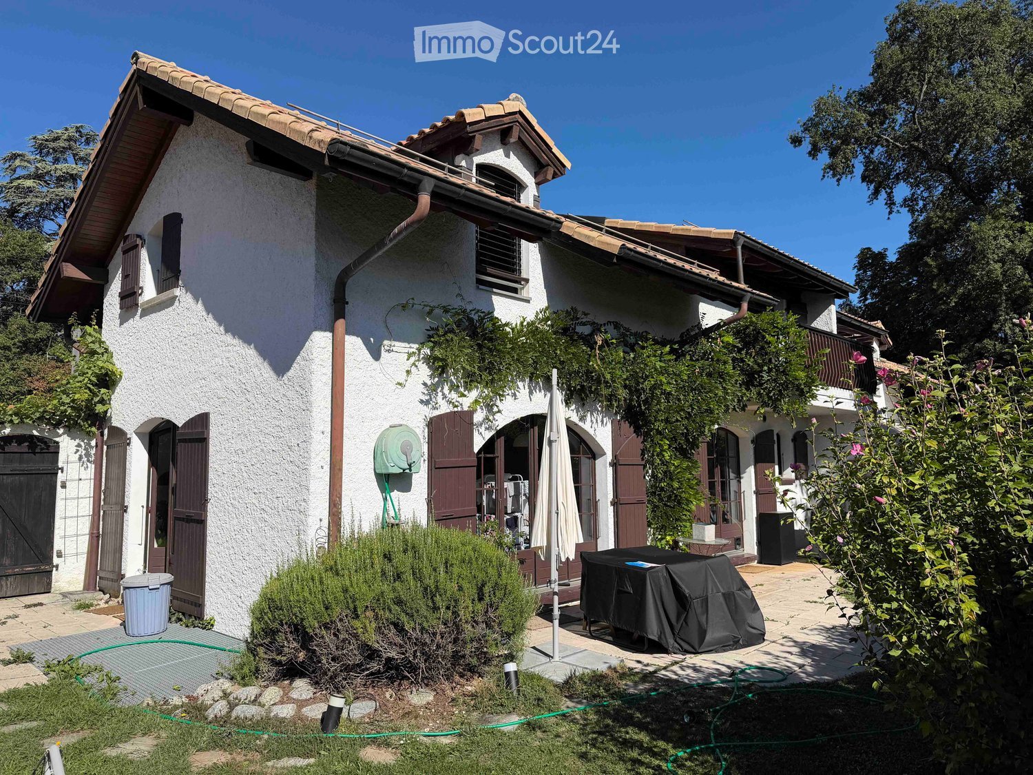 Two-story house with white stucco exterior, terracotta roof, multiple windows with shutters, and a covered patio area with outdoor furniture.