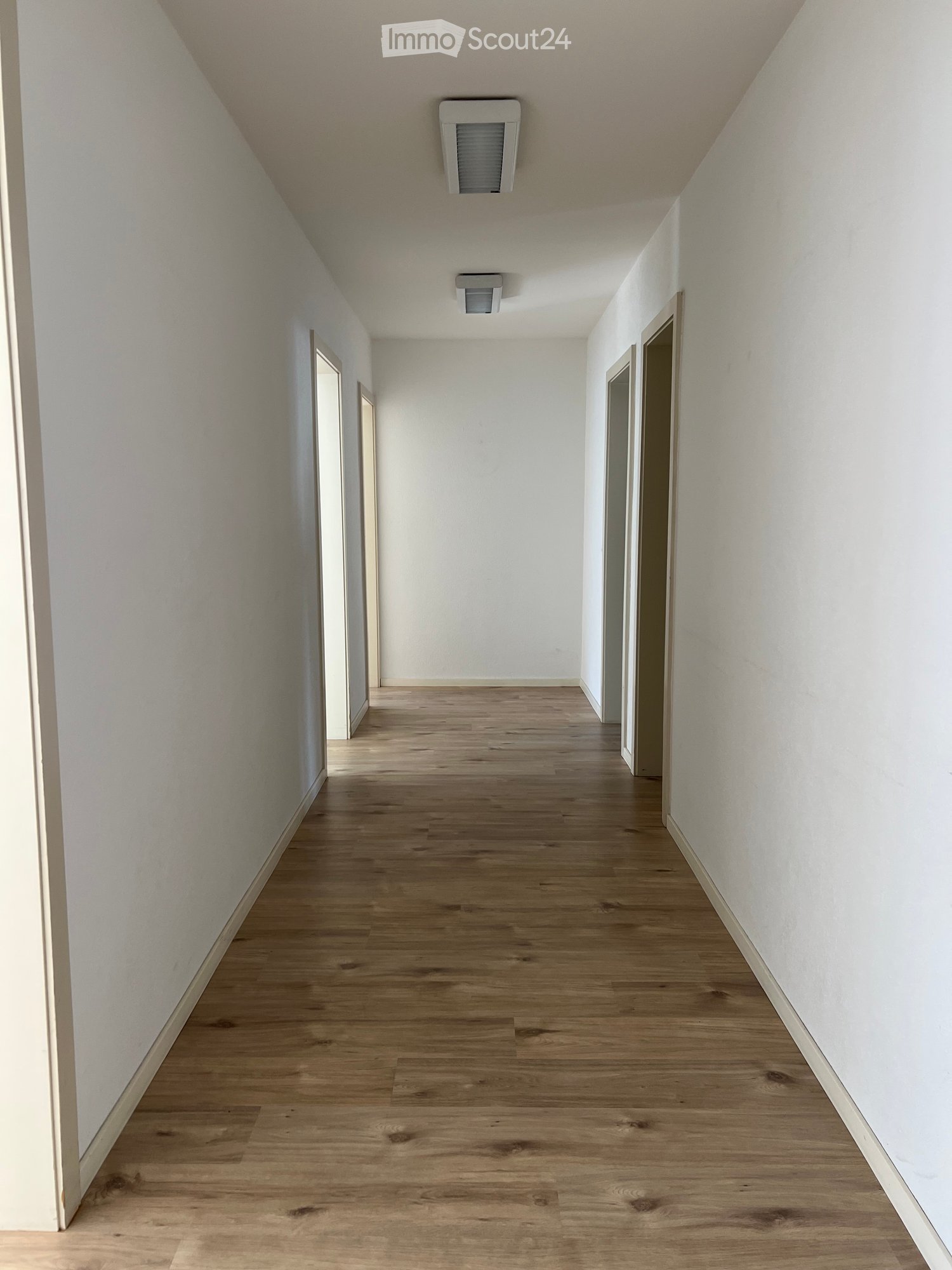 Empty hallway with light wooden flooring and white walls, featuring several doors and ceiling lights.