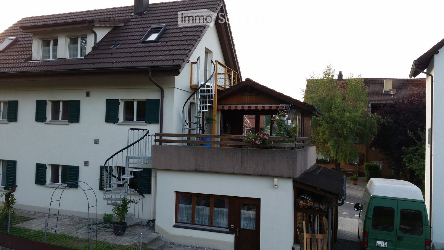 Two-story house with white exterior, green shutters, spiral staircase leading to a balcony, and a small garden area.