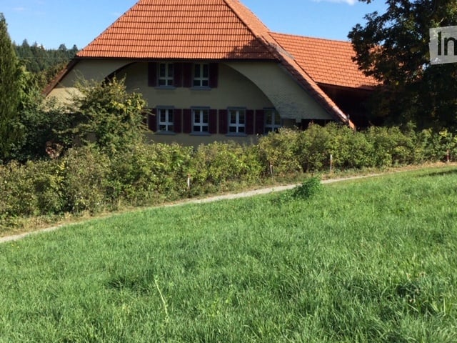 A two-story house with a red-tiled roof and beige walls, surrounded by greenery and a grassy lawn.