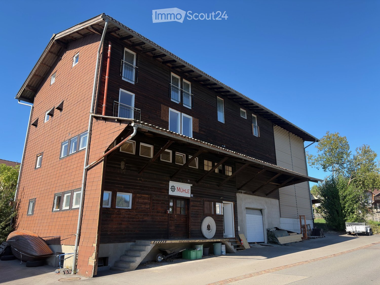 Two-story wooden building with brick and wood siding, multiple windows, balconies, and a sign reading "MÜHLE".