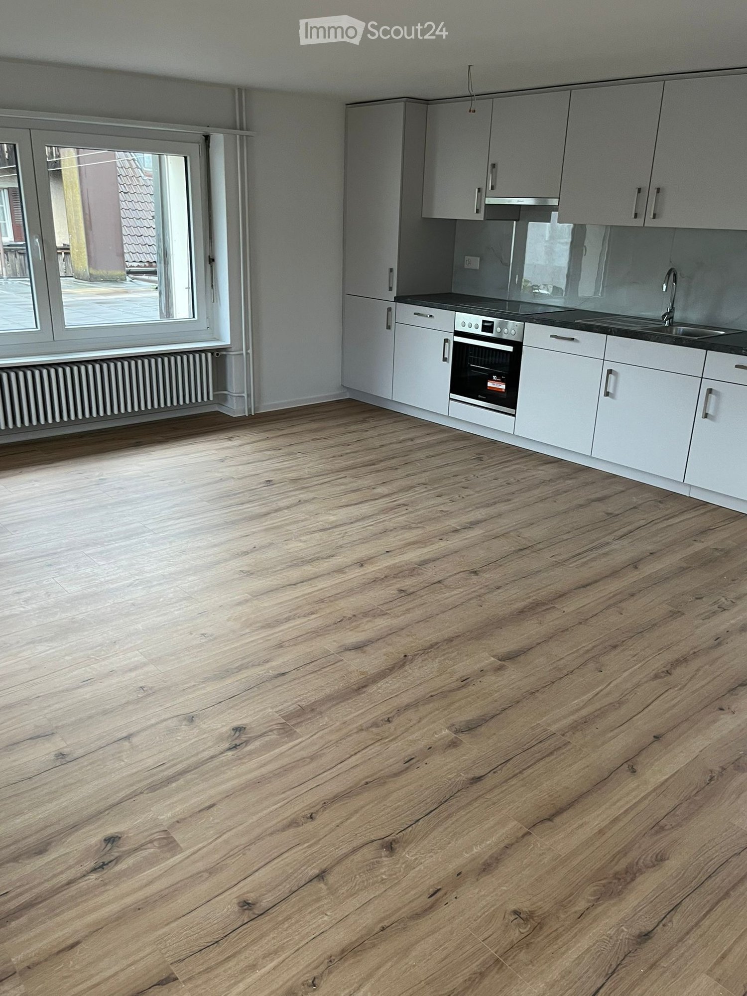 Empty kitchen with light wooden flooring, white cabinets, and a built-in oven. The space is unfurnished and features a large window with a view of the outside.