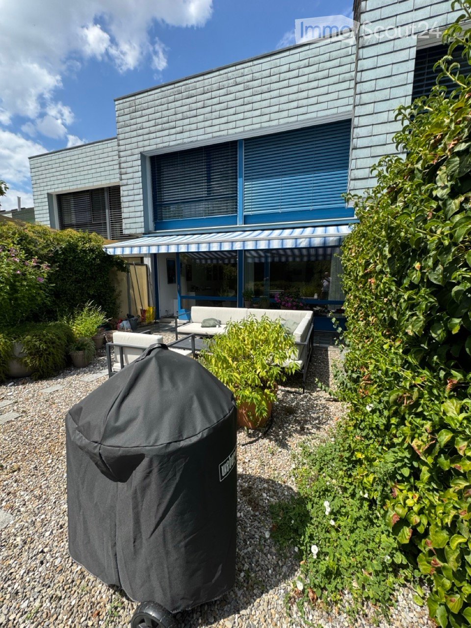 Outdoor patio area with a covered seating area, featuring a sofa, table, chairs, and a covered grill, surrounded by plants and a modern house with blue shutters.