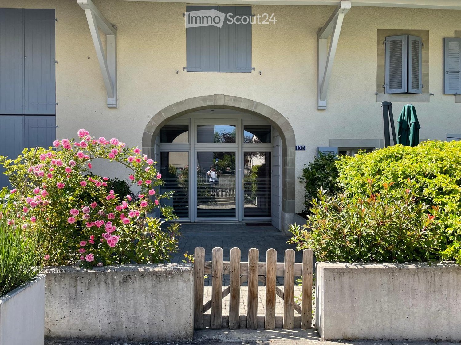 Two-story house with light yellow walls, grey shutters, arched entrance, and well-maintained garden with pink flowers and greenery.