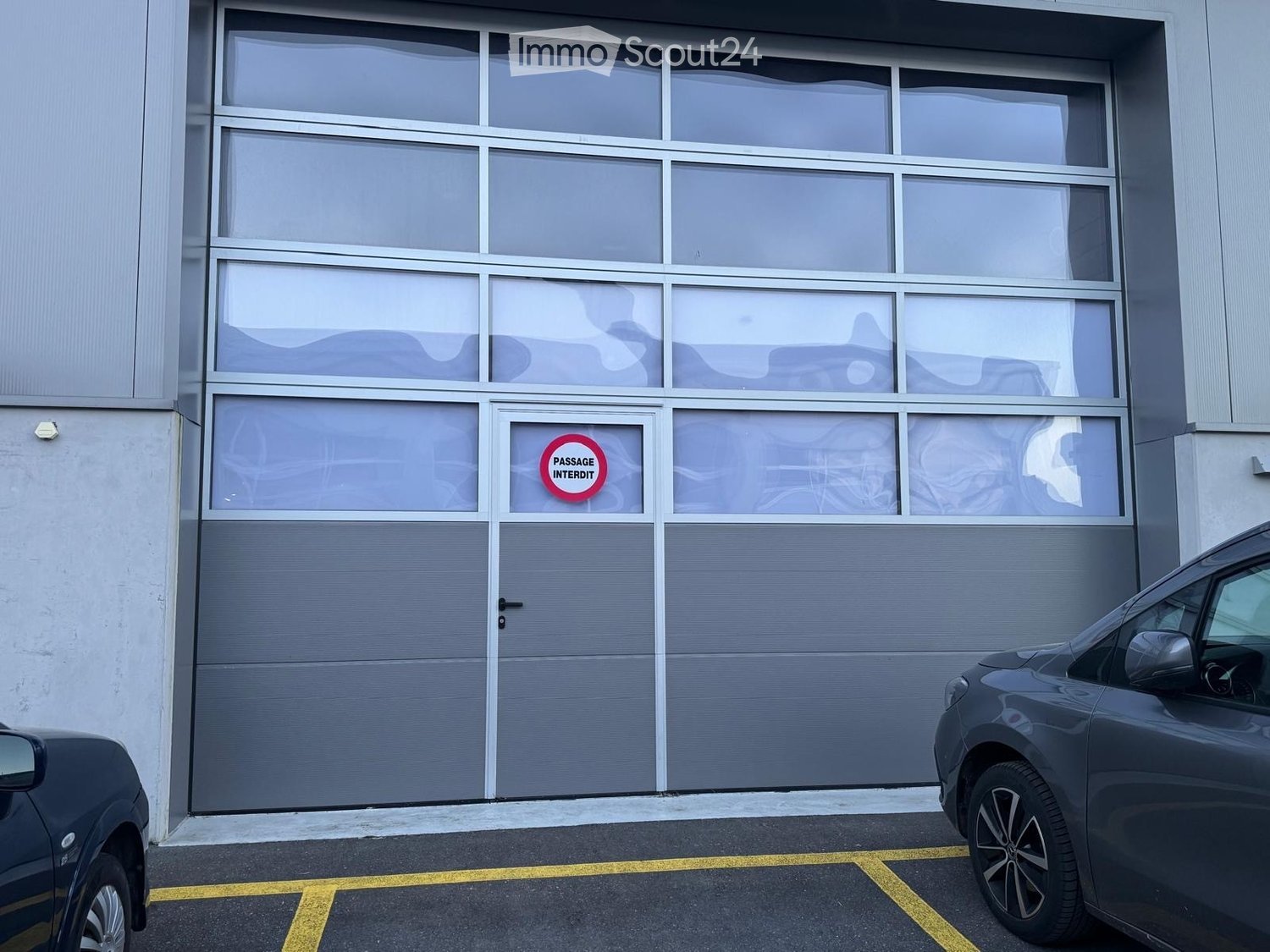 Gray building, large garage doors, 'Passage Interdit' sign, small pedestrian door, yellow parking lines