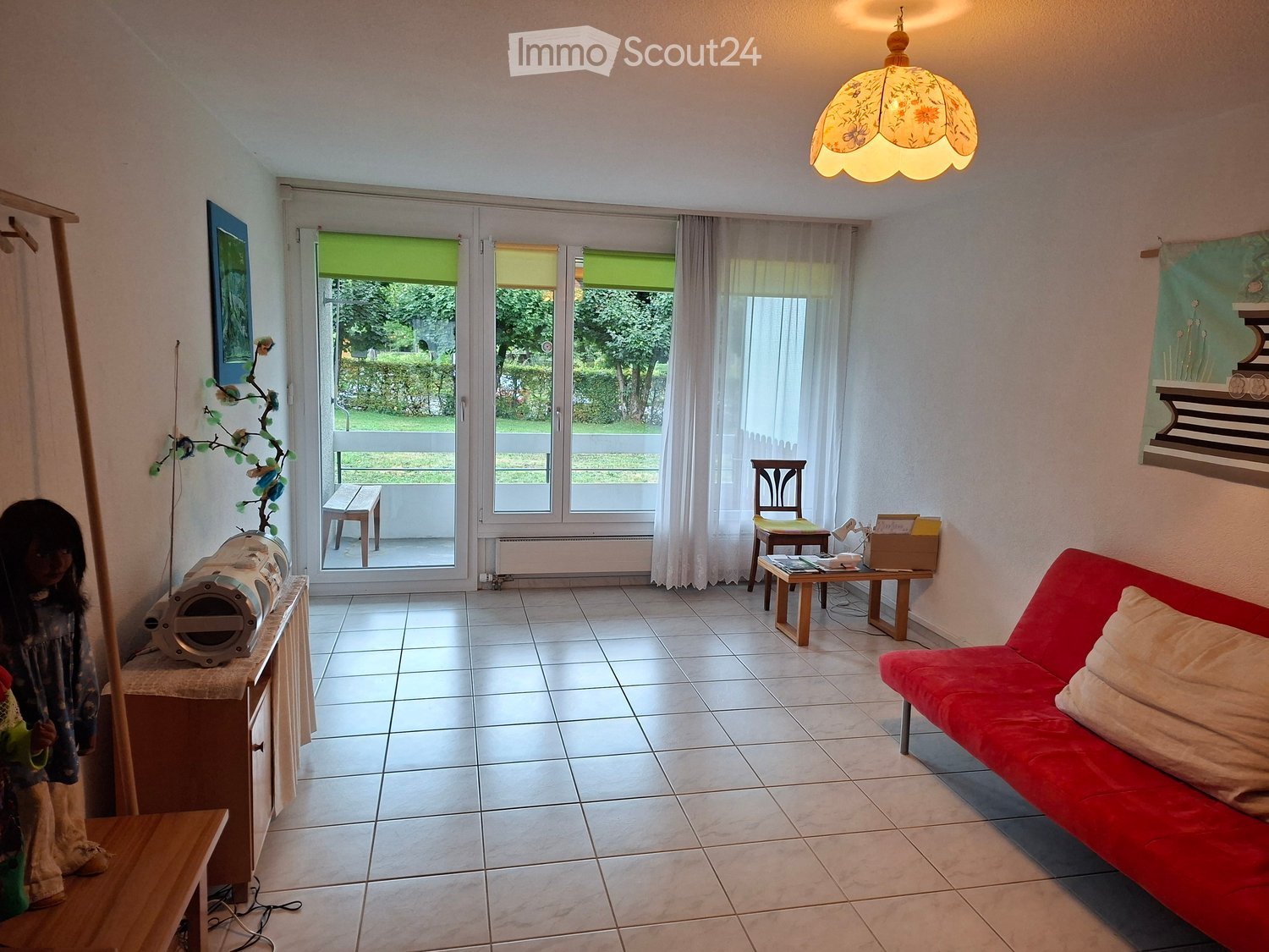 Bright living room with tiled floor, large windows, red sofa, small wooden table, and decorative items.