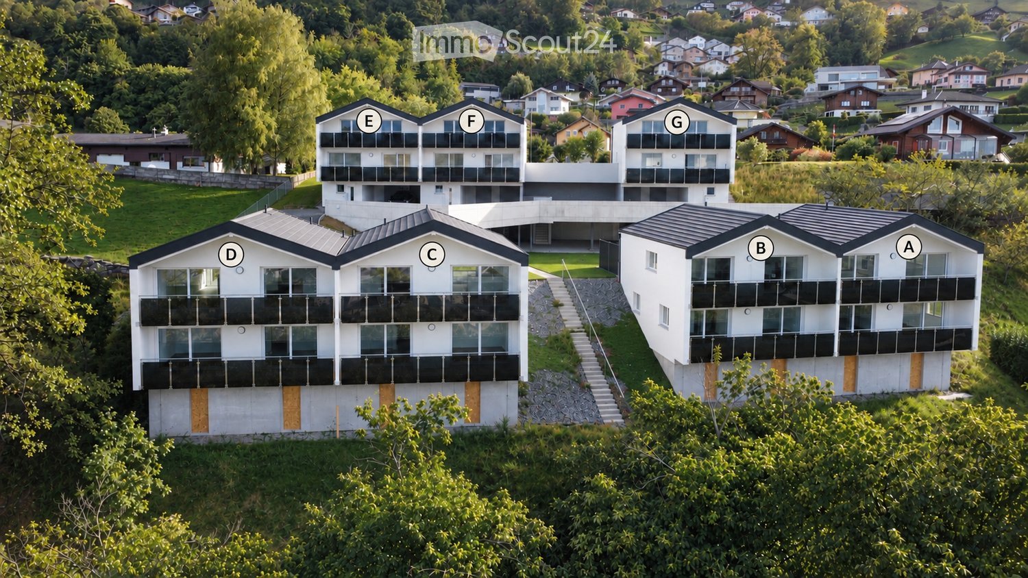 Aerial view of a modern residential complex with multiple buildings labeled A through G, featuring balconies, large windows, and a landscaped green area.