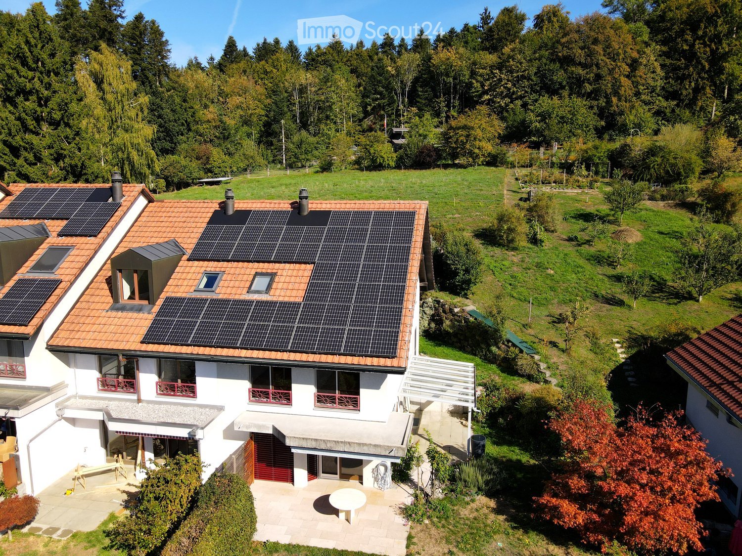 A two-story house with a red-tiled roof, solar panels, multiple windows, and a surrounding garden with trees and a small patio area.