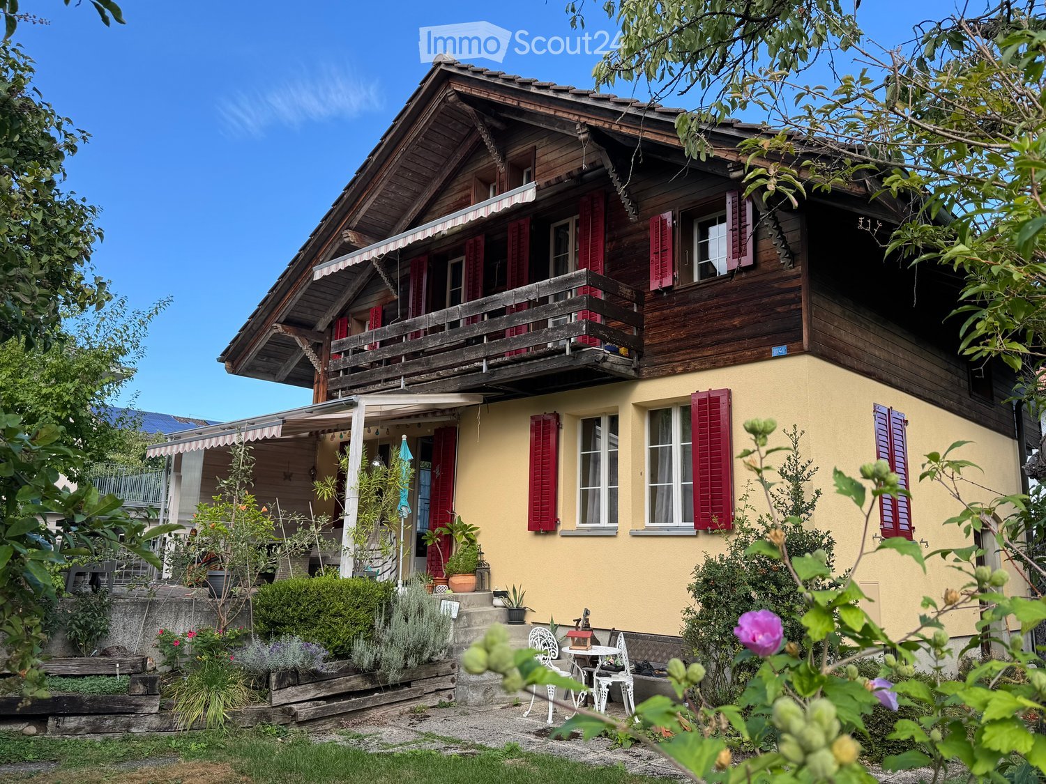 Two-story house with wooden exterior, red shutters, and a balcony on the upper level. The house features a covered entrance area with a small patio and outdoor seating, surrounded by greenery and plants.