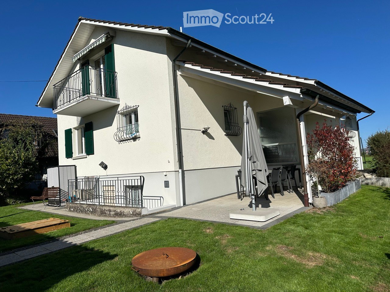 Two story house, white wall, brown roof, patio,  green shutters, wooden balcony, green grass, iron fence, table and chairs on patio