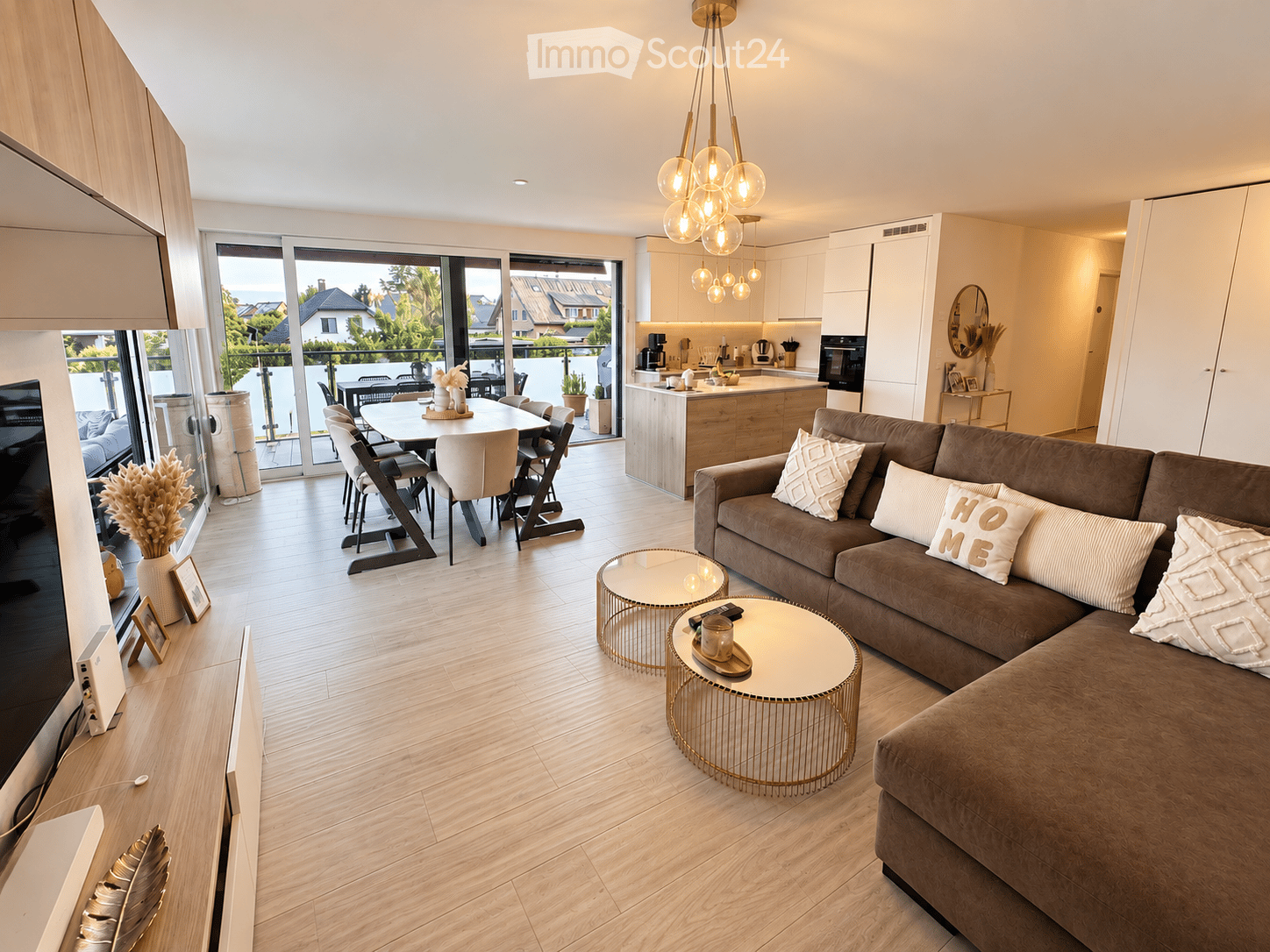 Modern living and dining area with a brown sectional sofa, coffee tables, dining table with chairs, and a kitchen area with cabinets and appliances. Large windows and sliding doors lead to a balcony.