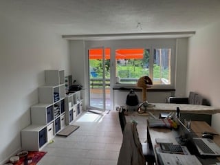 Bright, modern room with large window and sliding glass door leading to a balcony. Contains a desk with a laptop, chair, bookshelf, and a lamp.