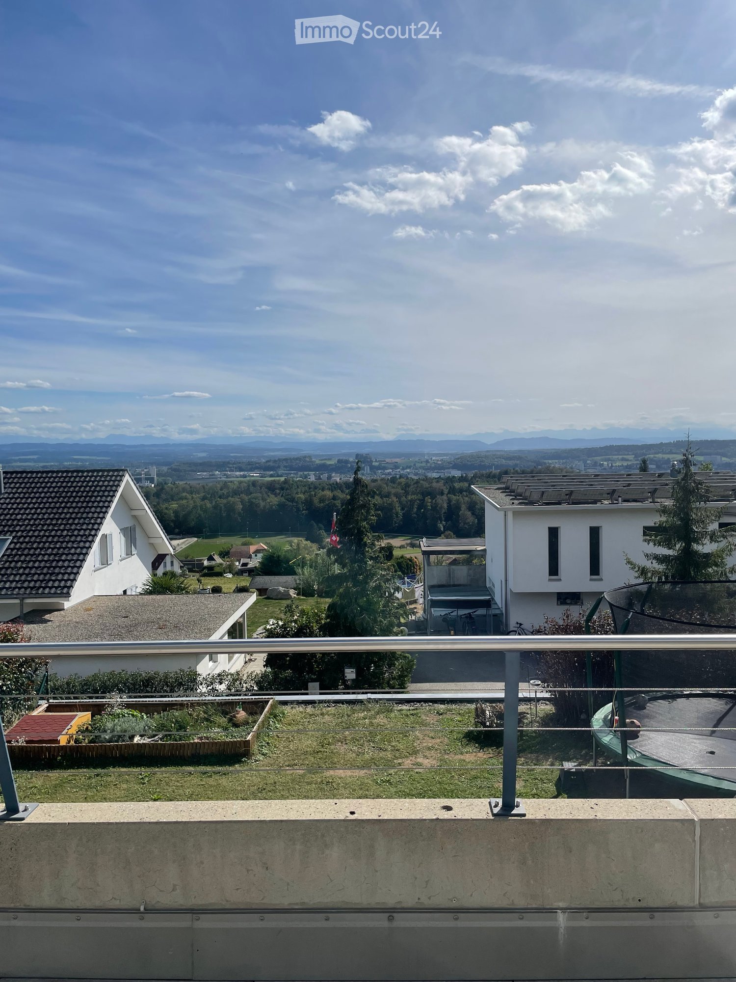 Balcony with metal railing overlooking a suburban area with houses, trees, and distant mountains under a partly cloudy sky.
