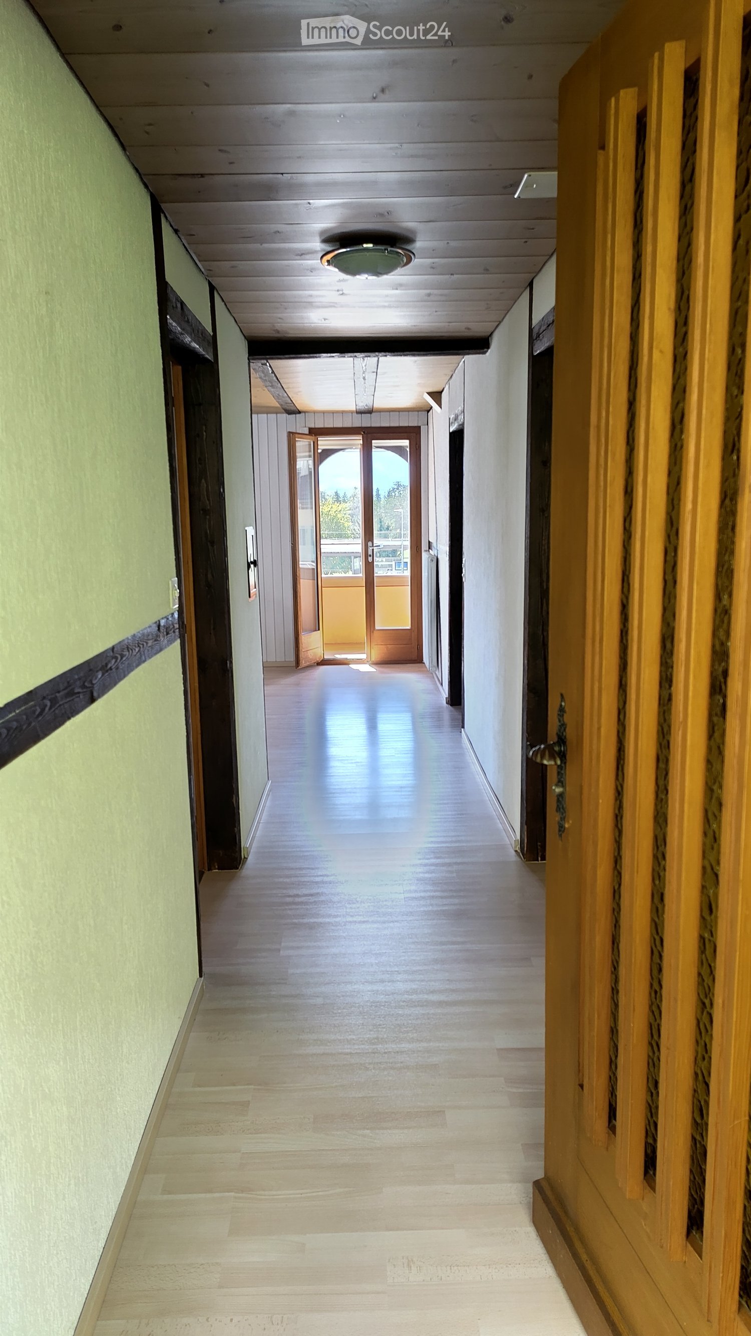 A long, well-lit hallway with wooden flooring, light-colored walls, and a ceiling light fixture. An open doorway at the end leads to another area with natural light.