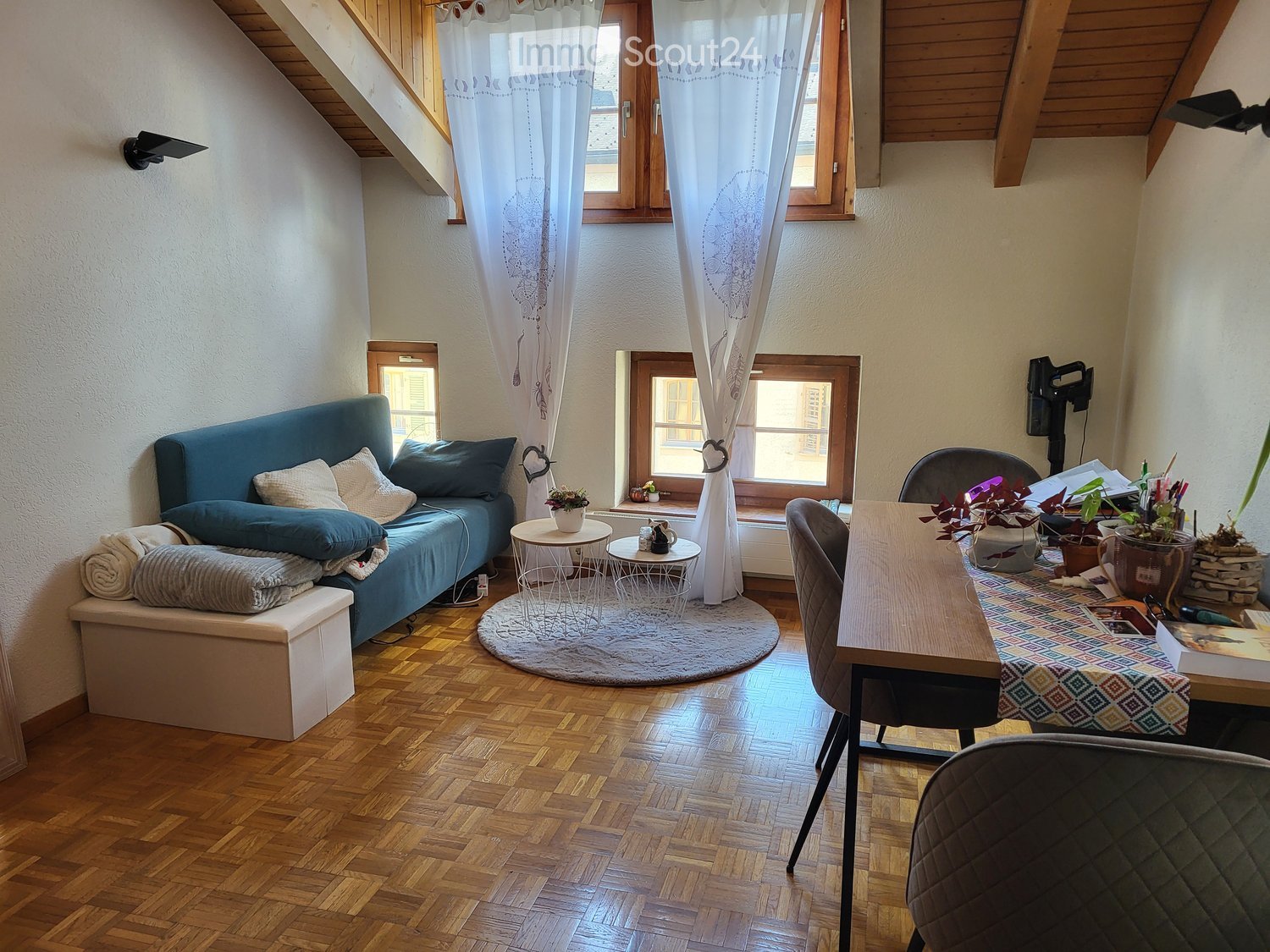 Cozy living room with wooden flooring, a blue sofa, small round tables, a desk with a chair, and decorative plants.